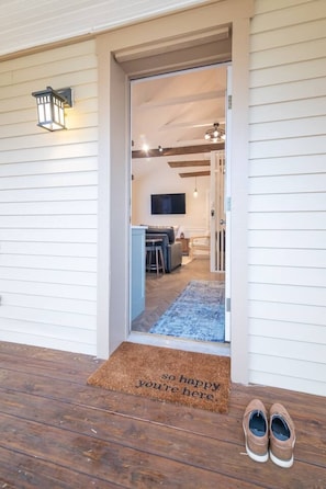 Um deck de madeira com um tapete na entrada, um par de sapatos e vista para uma sala de estar com televisão, sofá e mesa de jantar.