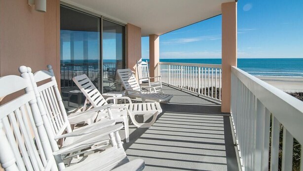 Un balcón con sillas y una mesa blancos, con vista a una playa.
