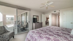 Un dormitorio con cama, sofá, televisor y puerta corrediza de vidrio que da a un balcón con mobiliario exterior.