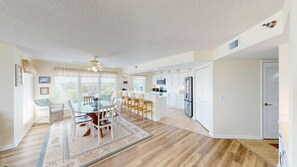 A spacious living area with a dining table, chairs, a kitchen with white cabinets, and a large window providing natural light. - Beachfront Haven 2BR Retreat Litchfield By the Sea (Pawleys Island)