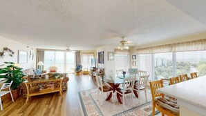 A spacious living room with a dining area, large windows, and a ceiling fan.