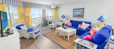 A living room with a blue sofa, white wicker chairs, a coffee table, and a view of the ocean through large windows.