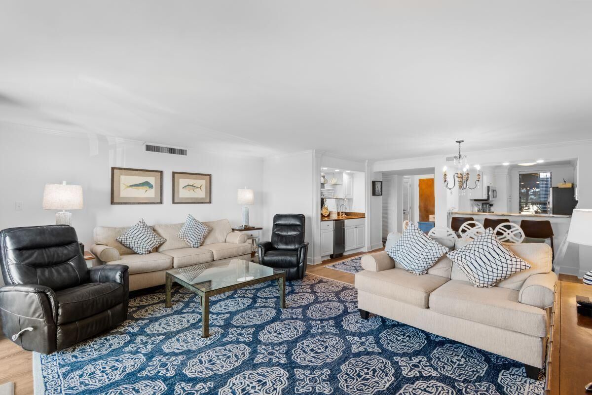 Een moderne woonkamer met een beige bank, een zwarte lederen fauteuil en een glazen salontafel. Er hangen twee ingelijste foto's aan de muur en er is een kroonluchter.