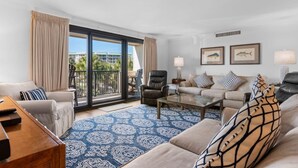 A modern living room with a large sliding glass door leading to a balcony, a sofa set, a coffee table, and two framed pictures on the wall.