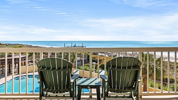 A balcony with two green Adirondack chairs and a small table, overlooking a pool and a beach.