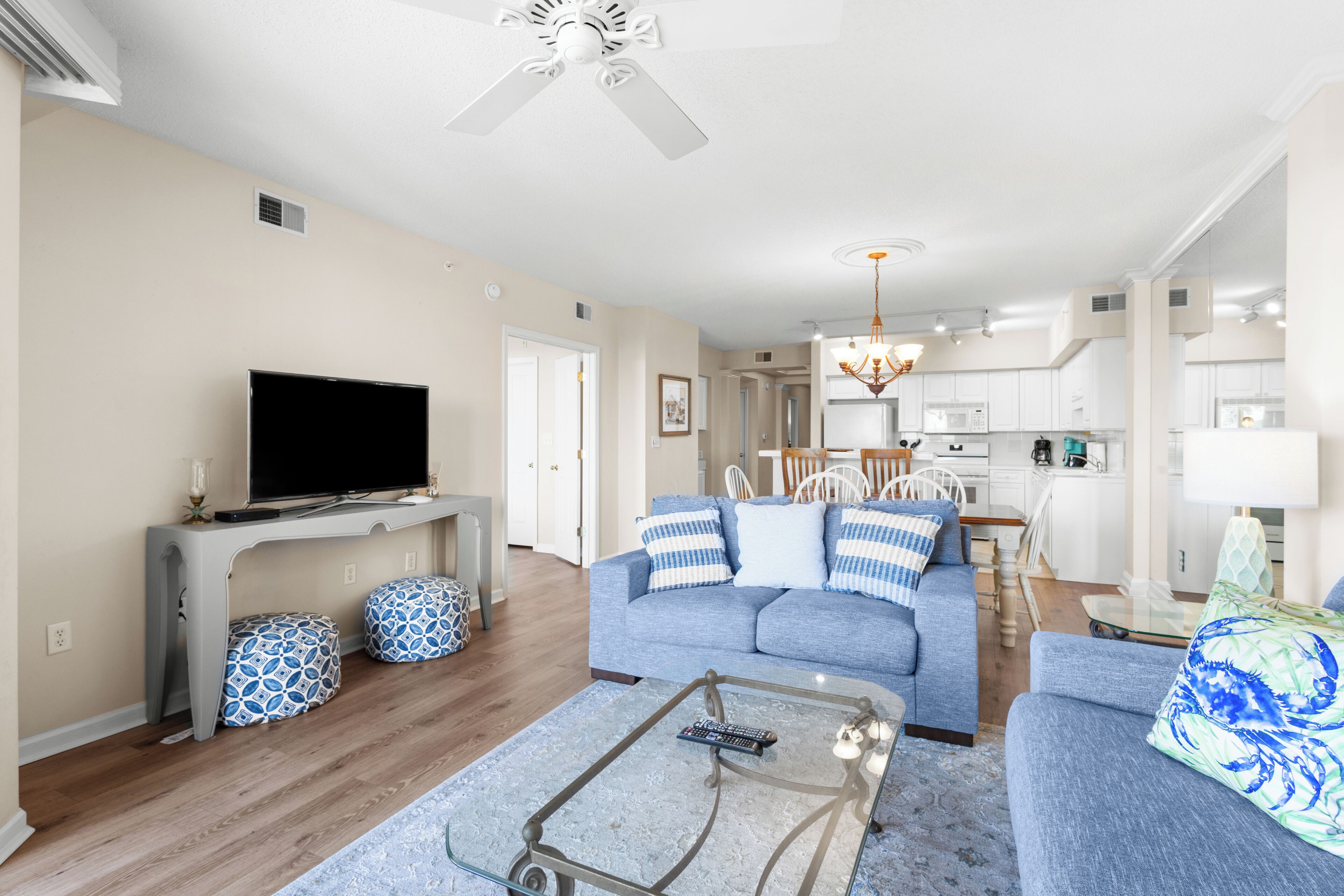 A living room with a flat-screen TV, a sofa, ottomans, a coffee table, and a ceiling fan.