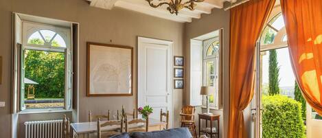 A room with a dining table, chairs, a sofa, a chandelier, and a large window with curtains.