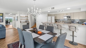 Une cuisine moderne avec des meubles blancs, un îlot central doté de tabourets, un lustre et un coin repas avec une table et des chaises.