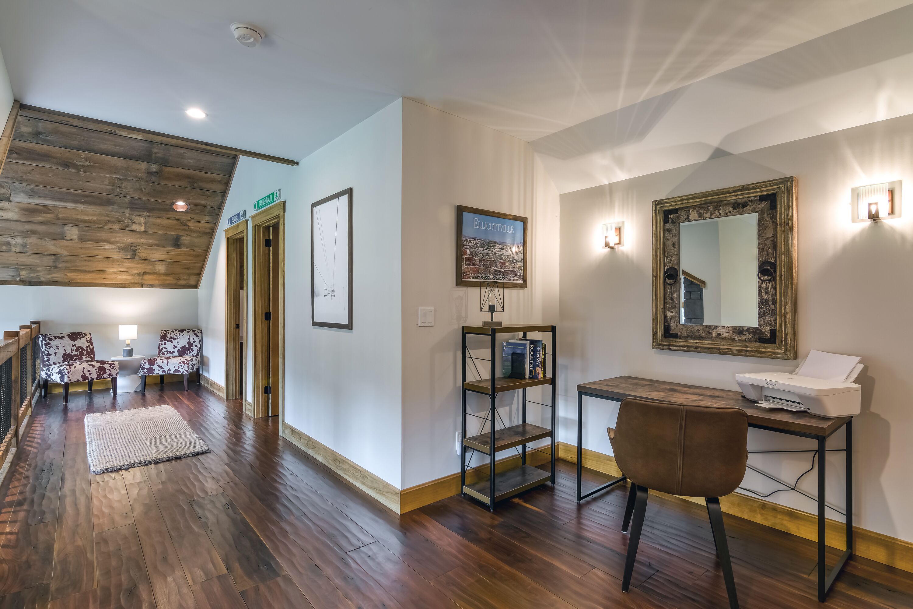 A spacious room with a wooden ceiling, a desk with a printer, two chairs, a rug, and framed artwork on the walls.