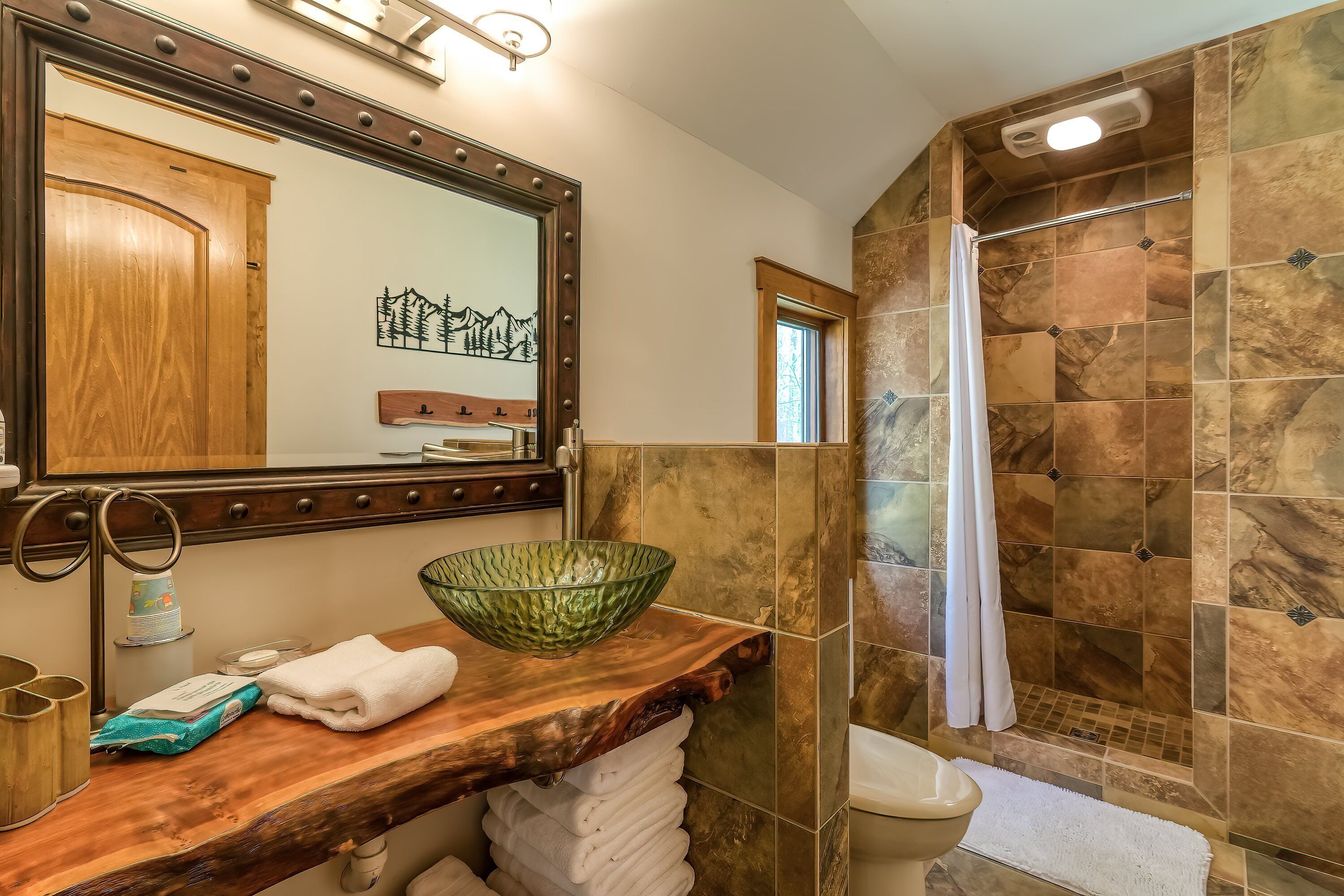 A bathroom with a wooden vanity, a green glass bowl, a towel rack, and a shower with stone tiles.