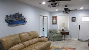 A living room with a tan leather couch, a green armchair, a small table with a lamp, and a ceiling fan. - Bayou Living (Barataria)
