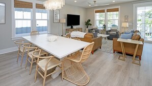 A spacious living room with a large white dining table, wicker chairs, a sofa, and a television.