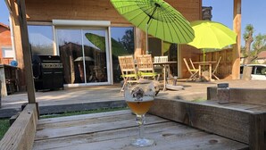 A glass of beer on a wooden bench with a patio area, outdoor furniture, and a barbecue grill in the background.