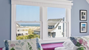 A bedroom with a large window overlooking a beach and houses, a bed with floral pillows, and a framed picture on the wall.
