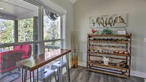 Una habitación con una mesa de madera y taburetes de bar, un jarrón de vidrio con ramas, una estantería con copas y jarras de cobre, y un cuadro de pájaros en la pared.