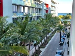Un hotel moderno con una fila de palmeras a lo largo de la calle, vista despejada del océano y un estacionamiento con coches.