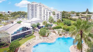 A hotel with a swimming pool, surrounded by greenery and a large building in the background.