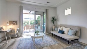 A living room with a sofa, a coffee table, a desk, and a chair. There is a window with curtains, a plant, and a ceiling fan.