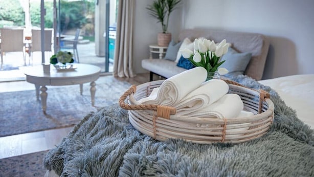 A cozy room with a wicker basket of towels, a plush rug, a sofa with a plant, and a view of the outdoors.