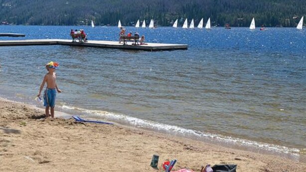 Ein Strand mit einer Anlegestelle, Segelbooten und Bergen im Hintergrund.