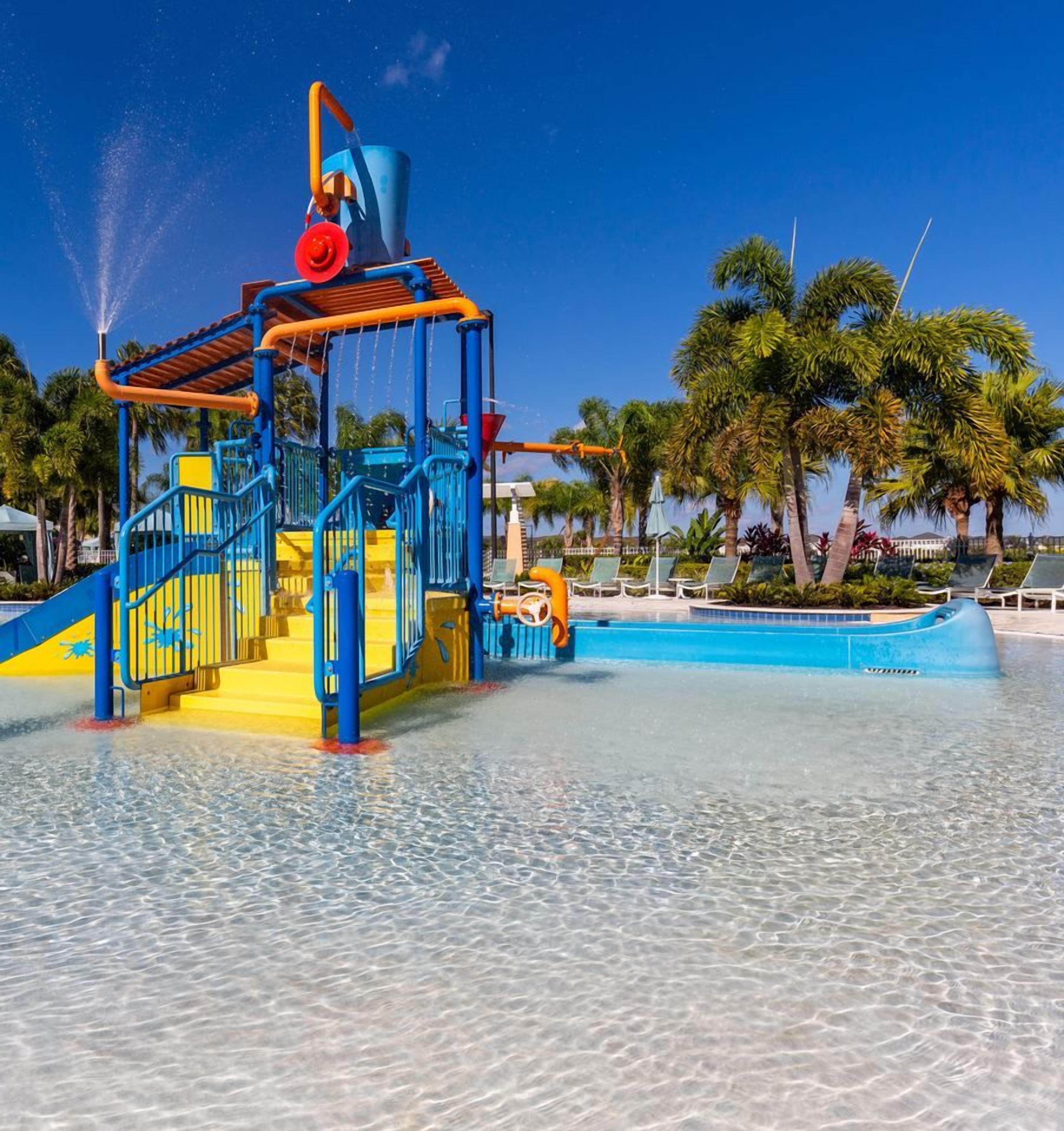 Ein Wasserspielbereich mit Rutsche und Wasserstrahl, umgeben von einem Schwimmbecken und Palmen.