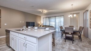 Una cocina moderna con una isla central, un comedor con mesa y sillas, y una sala de estar con sofá y televisor.