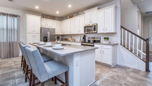 Una cocina moderna con armarios blancos, una isla central con taburetes y electrodomésticos como microondas y refrigerador.
