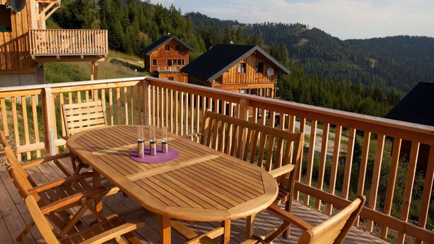 A wooden deck with a round table and chairs, overlooking a forested mountain area.
