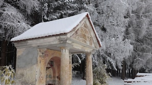 A snow-covered gazebo with a tiled roof and stone columns, surrounded by trees and snow.