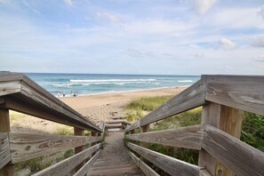 Un escalier en bois mène à une plage d’où l’on a une vue dégagée sur l’océan et où l’on aperçoit des personnes au loin.