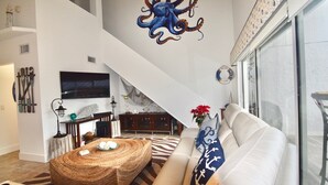 Un ampio soggiorno con soffitto alto, un grande murale a forma di polpo, un divano confortevole, un tavolino da caffè in fibra naturale e una scala che conduce a un livello superiore.