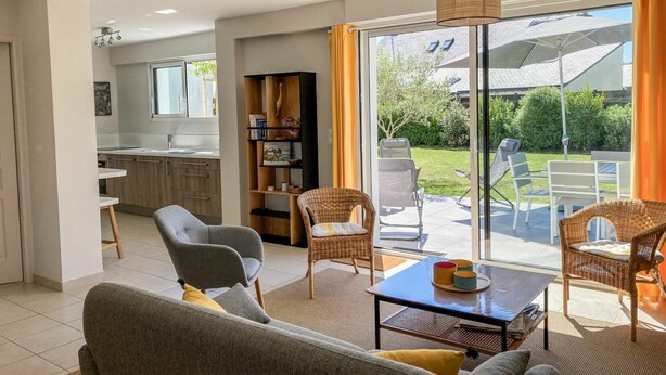 Ein modernes Wohnzimmer mit einer Couch, Sesseln und einem Tisch. Im Hintergrund ist eine Küche mit einem Fenster und einem Bücherregal zu sehen.