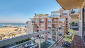 Un balcon avec vue sur la plage et l’océan, un espace de stationnement avec des voitures, et un immeuble doté de nombreux balcons et fenêtres.