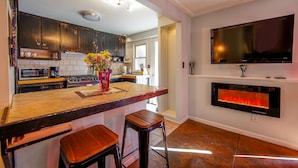 A modern kitchen with dark cabinets, a large island with stools, a flat-screen TV, and a built-in fireplace.