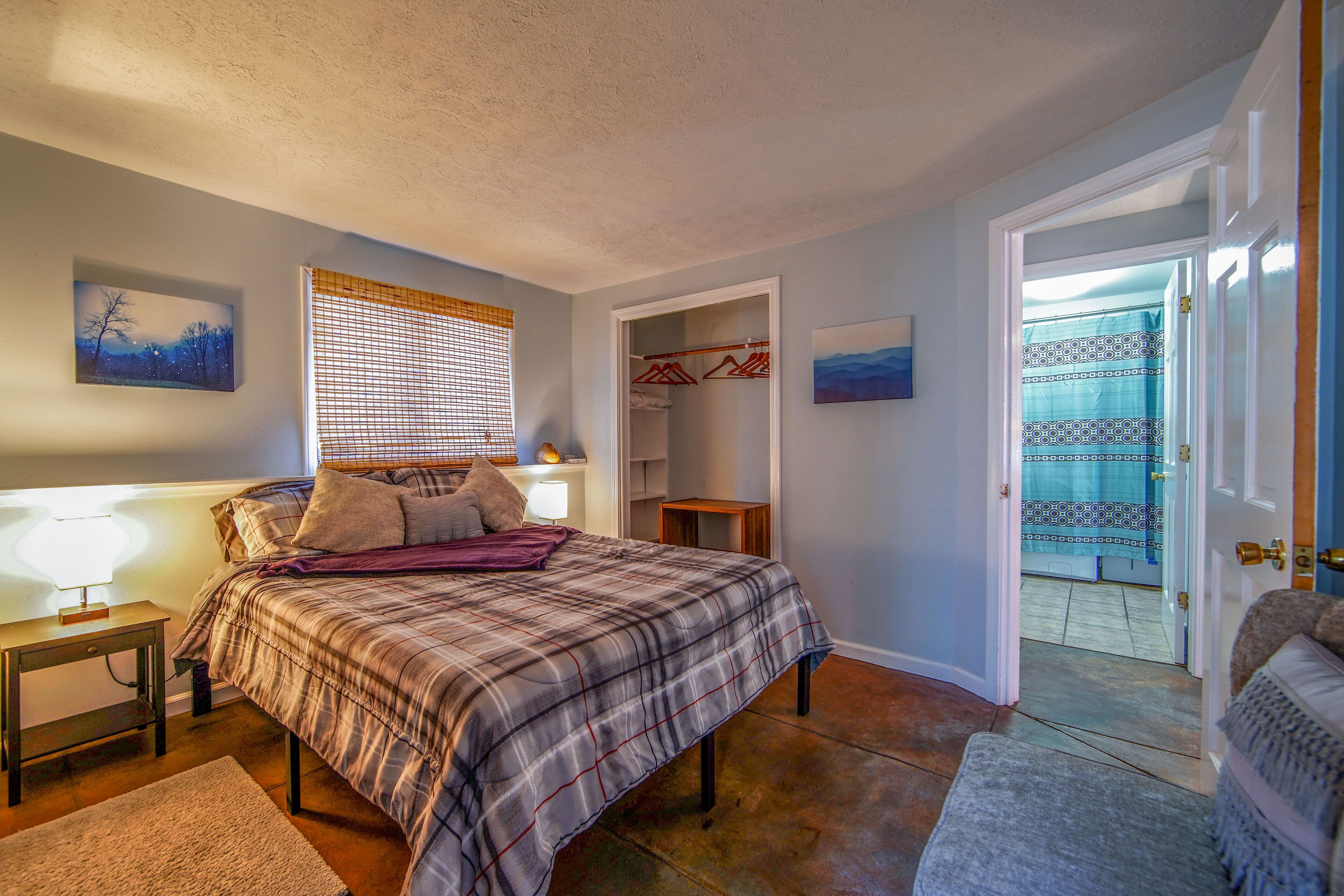 A bedroom with a bed, bedside table, and a nightstand. There is a window with blinds, a door leading to a bathroom, and a hallway.