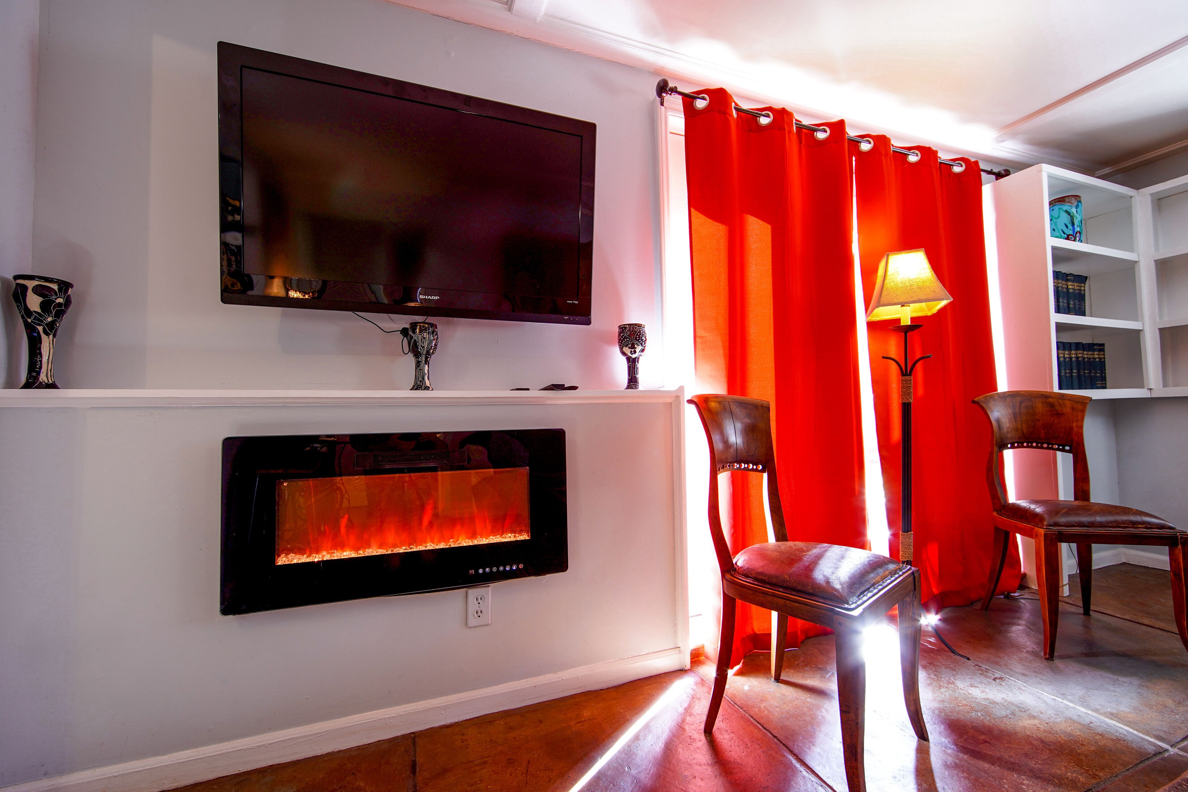 A living room with a flat-screen TV mounted above a fireplace, two chairs, a red curtain, and a floor lamp.
