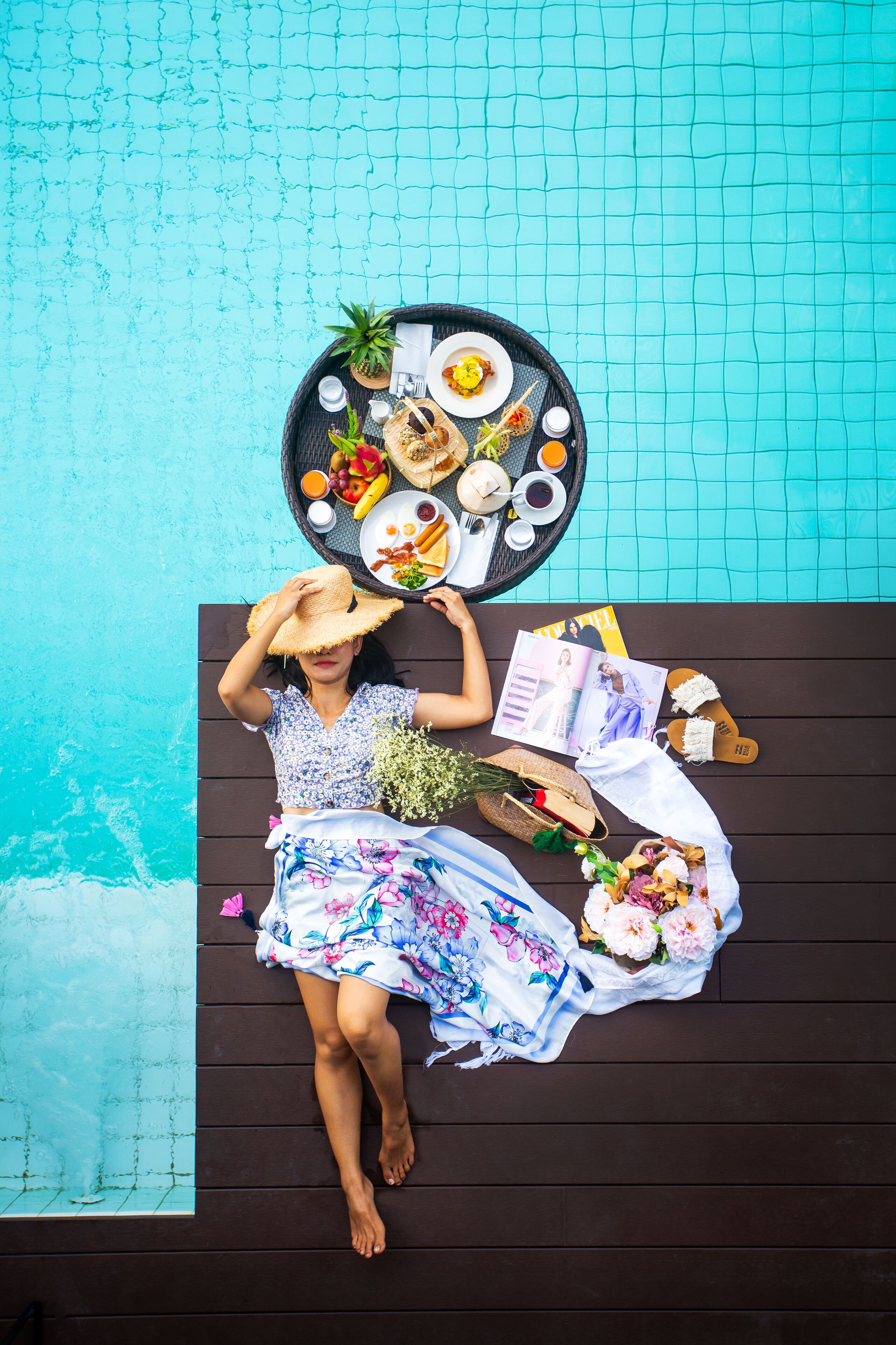 A person lying on a wooden deck by a pool with a tray of food, a magazine, and a hat.