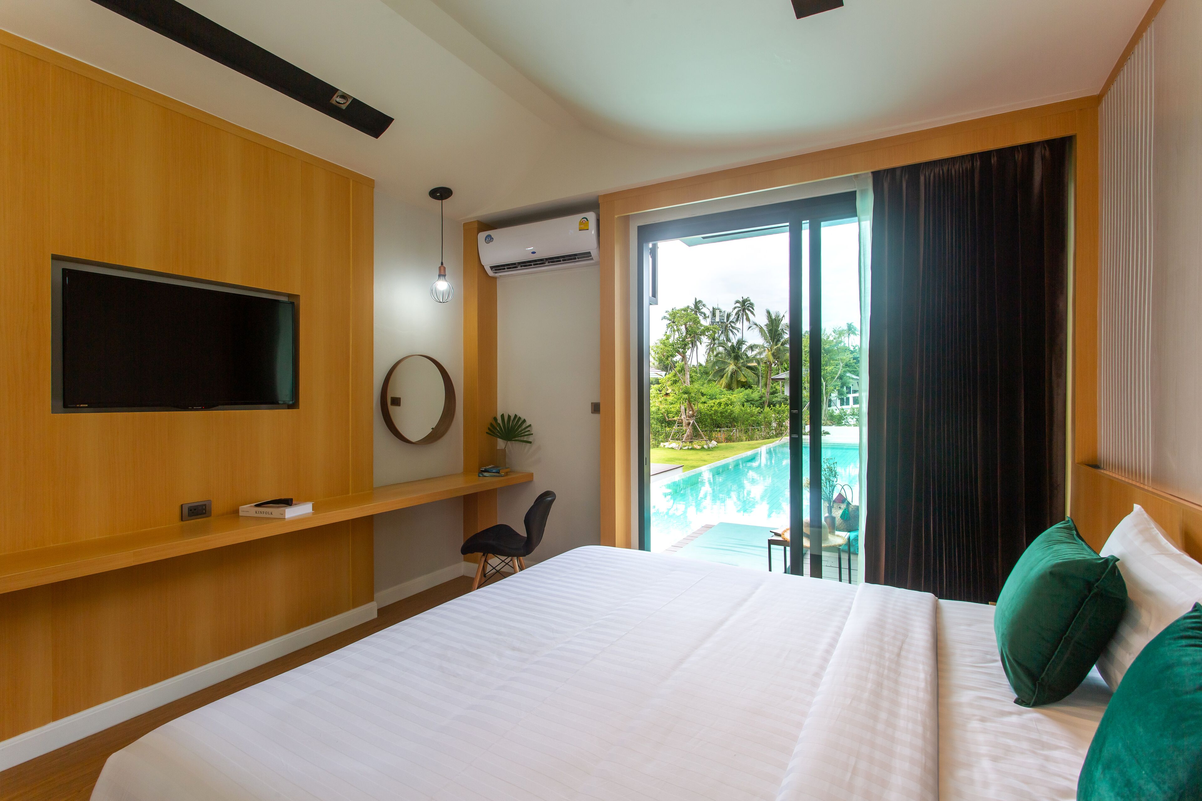 A modern hotel room with a large bed, a flat-screen TV mounted on the wall, a small table with a chair, and a view of a swimming pool through a glass door.