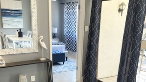 Un salon avec un canapé bleu foncé, un coussin à motifs, un vase blanc et un miroir au mur. Un couloir mène à une chambre à coucher où se trouve un rideau à motifs.