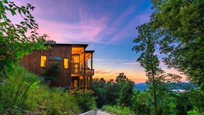 A modern two-story wooden house with a balcony, surrounded by lush greenery and a concrete pathway. - Capstone - Aska Adventure Area (Blue Ridge)