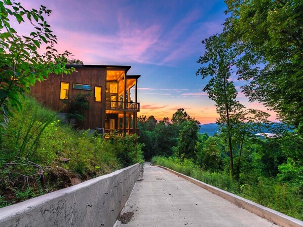 A modern two-story wooden house with a balcony, surrounded by lush greenery and a concrete pathway.