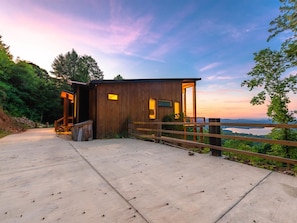 A modern wooden cabin with a concrete deck, surrounded by lush greenery and a scenic view.