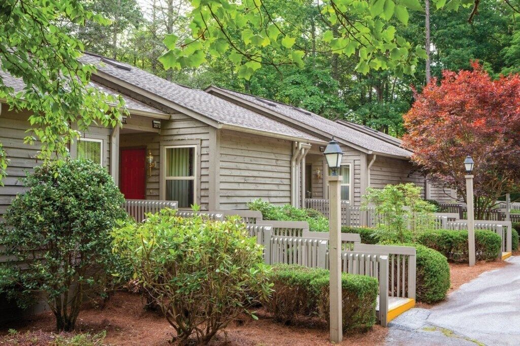Ein Bungalow mit roter Tür, umgeben von einem Holzzaun und einem kleinen Garten mit verschiedenen Sträuchern und einem Baum mit Herbstlaub.