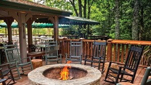 Un espace extérieur aménagé en terrasse, avec un foyer en pierre servant de coin salon, des tables et des chaises en bois, le tout entouré d’arbres.