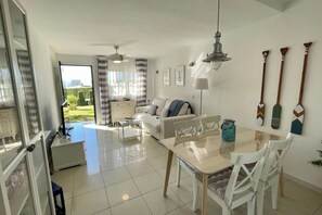 Een moderne woonkamer met een bank, een eettafel en stoelen. Er is een plafondventilator, een lamp en een raam met gordijnen.