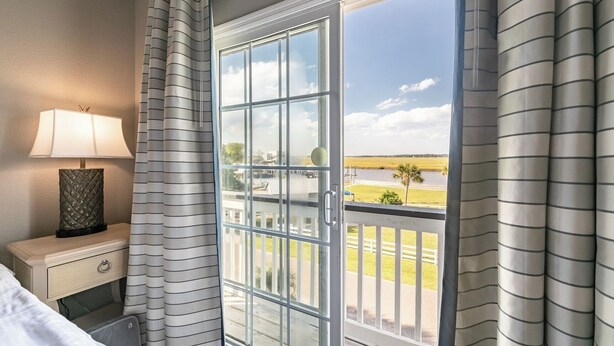 Una habitación de hotel con una cama, una mesita de noche con una lámpara, una ventana con vista a una playa y una valla.