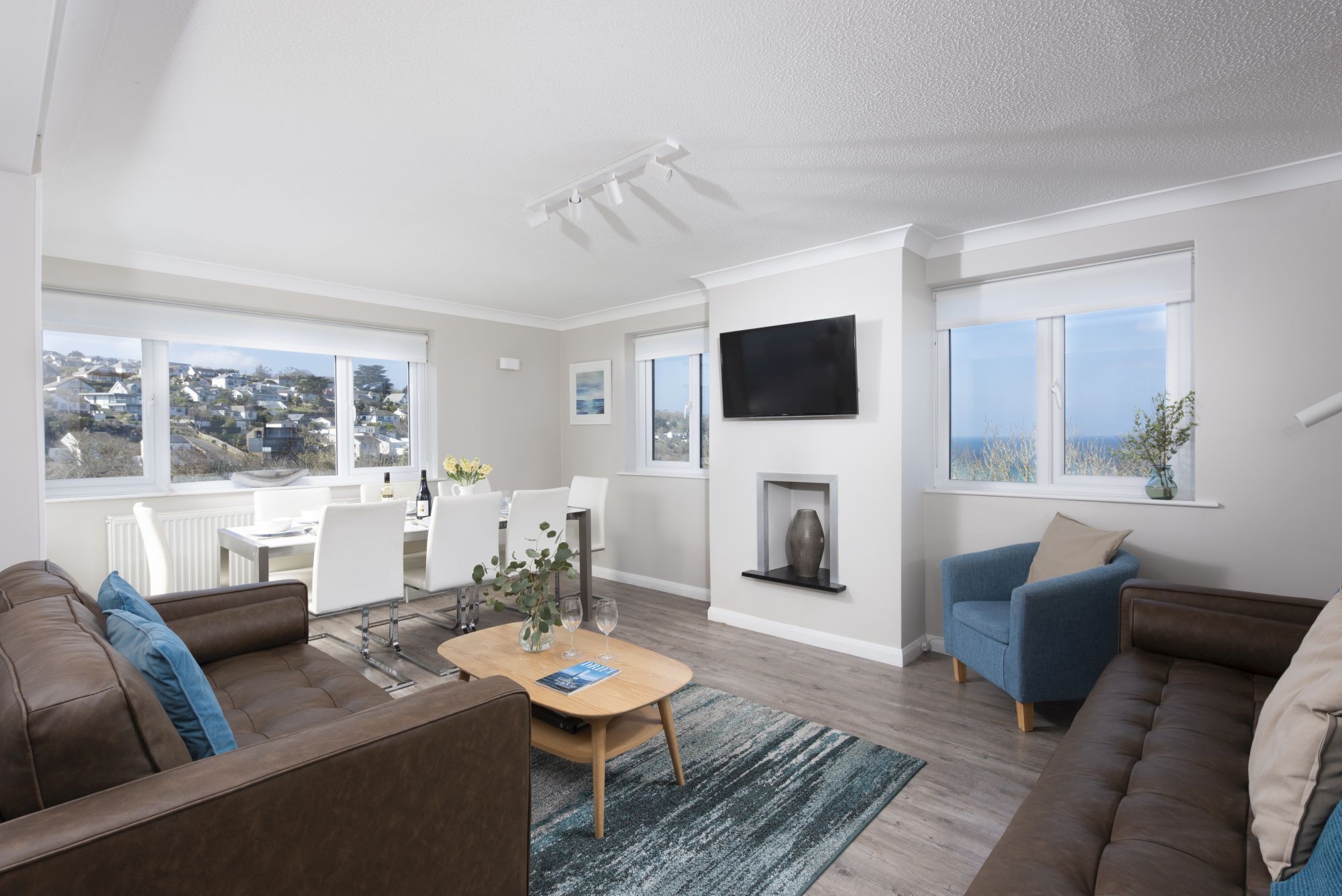 A modern living room with a sofa, a coffee table, and a TV mounted on the wall. There are windows providing views of the outside.
