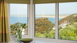 Una ventana con vista al mar y colinas, un florero con flores, y un cuenco en el alféizar.