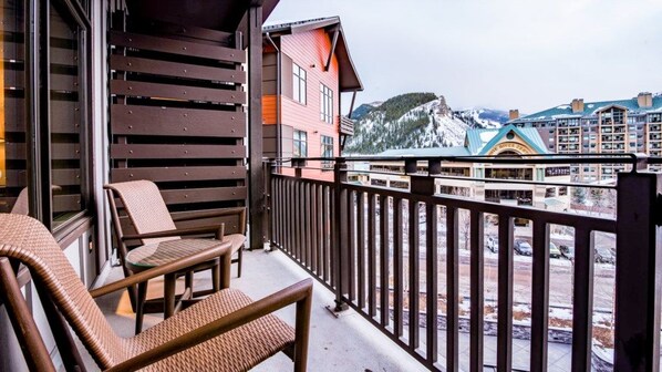 A balcony with wicker chairs, a wooden railing, and a view of a snow-covered mountain and buildings. - Avon Colorado | Couples Ski Retreat | Hot Tub + Ski (Avon)