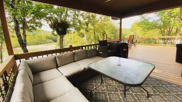 Ein überdachter Außenbereich mit einer Couch und einem Tisch, umgeben von einer Holzterrasse und Bäumen.